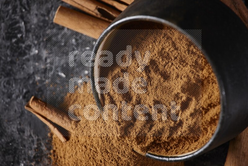 Black pottery bowl over filled with cinnamon powder and cinnamon sticks around the bowl on a textured black background
