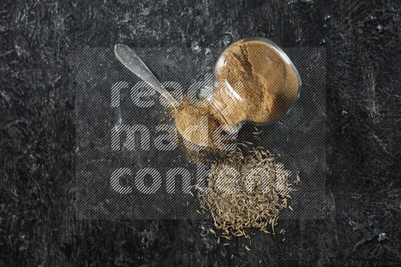 A flipped glass spice jar and a metal spoon full of cumin powder and powder spilled out with cumin seeds on a textured black flooring