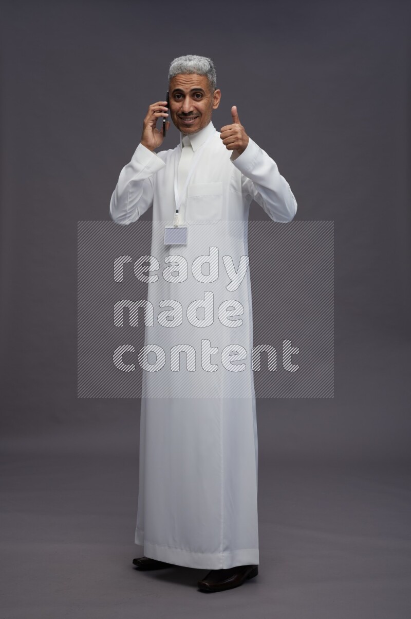 Saudi man wearing thob with neck strap employee badge standing talking on phone on gray background