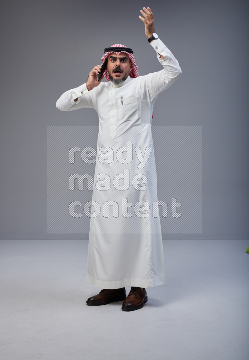 Saudi man Wearing Thob and red Shomag standing talking on phone on Gray background