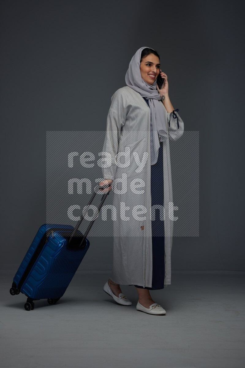 A Saudi woman wearing a light gray Abaya and head scarf standing and talking in the phone while holding shopping bags eye level on a grey background