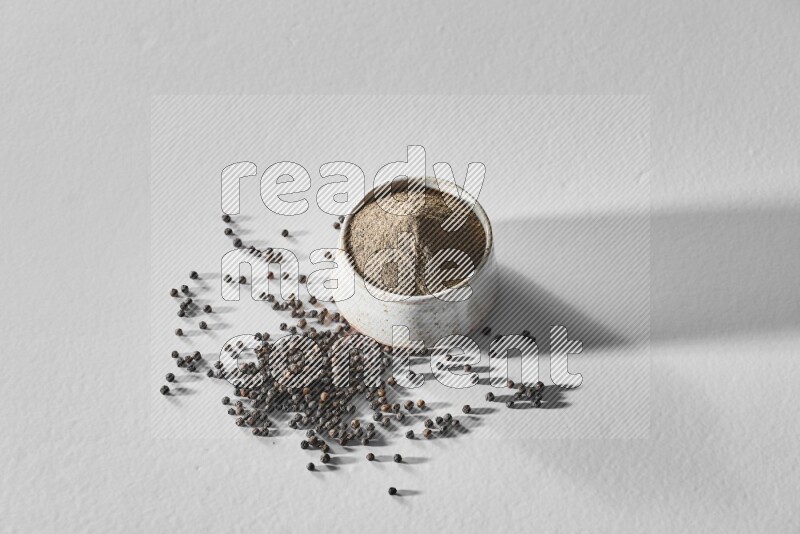 A white ceramic bowl full of black pepper powder and black pepper beads spread on white flooring