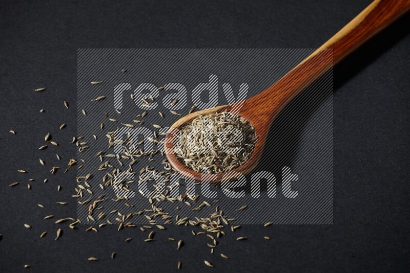 A wooden ladle full of cumin seeds on black flooring