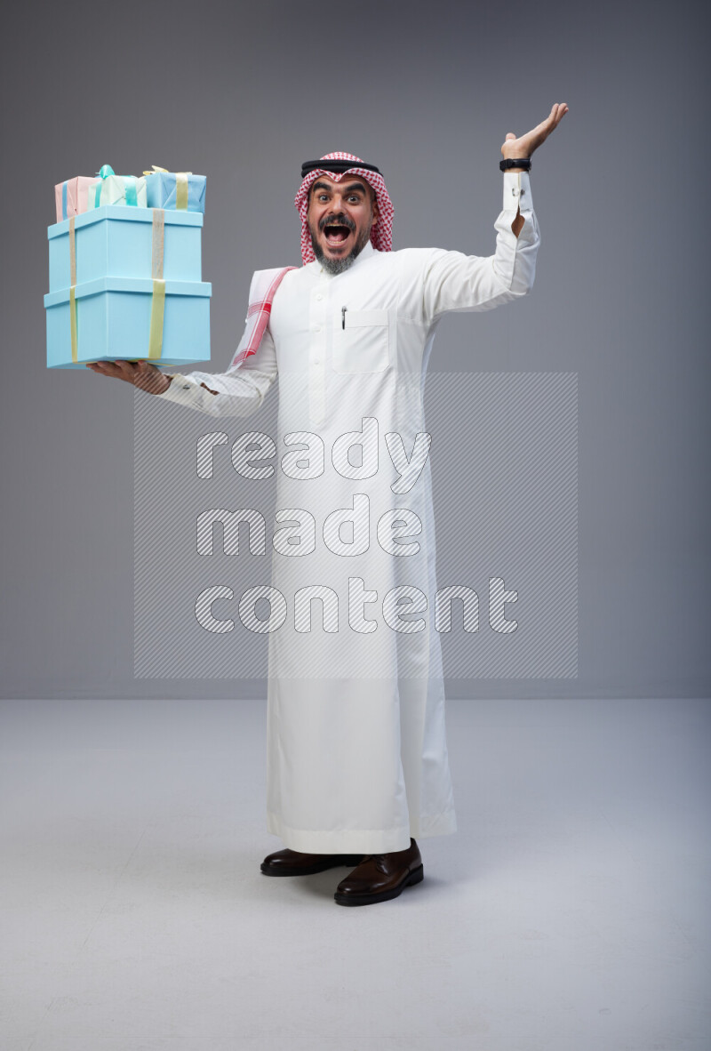 Saudi man Wearing Thob and red Shomag standing holding gift box on Gray background