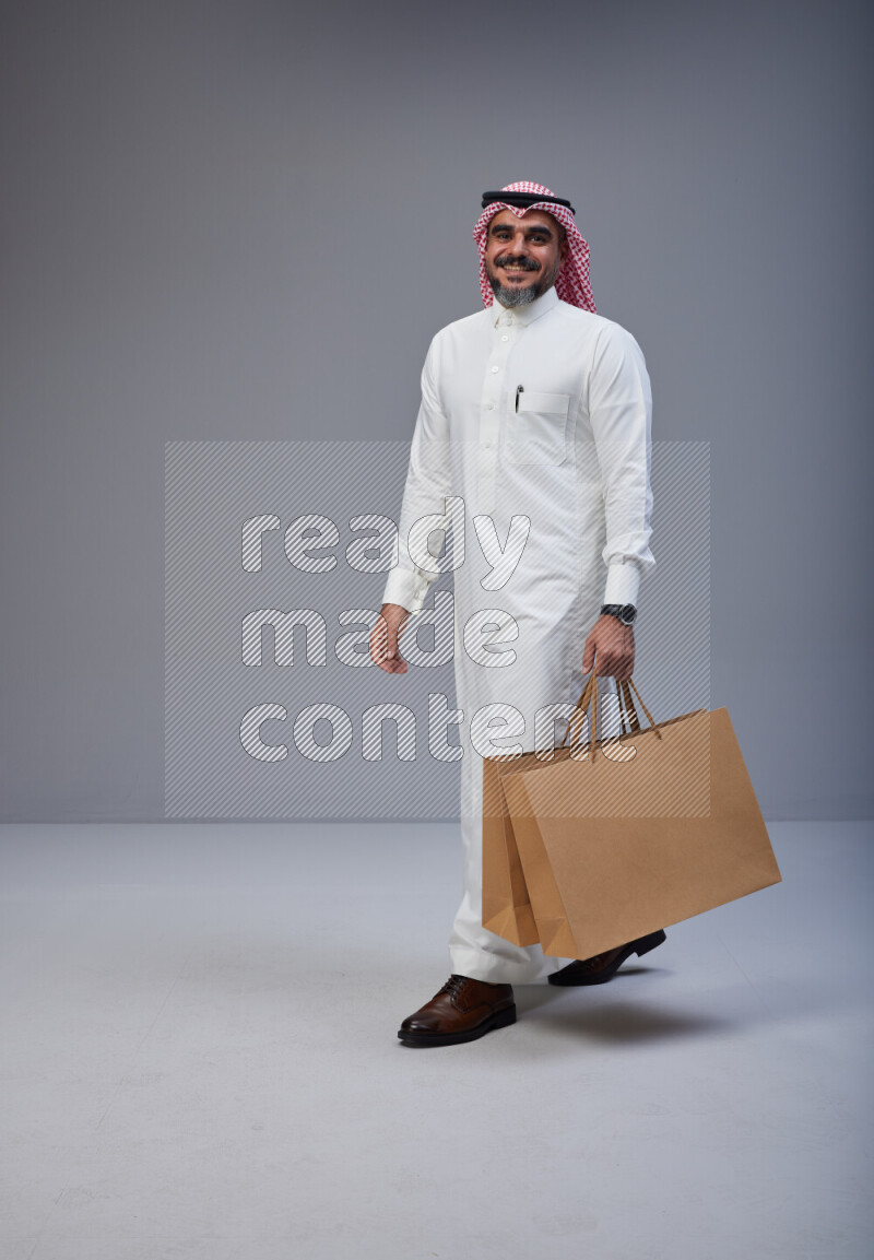 Saudi man Wearing Thob and red Shomag standing holding shopping bag on Gray background