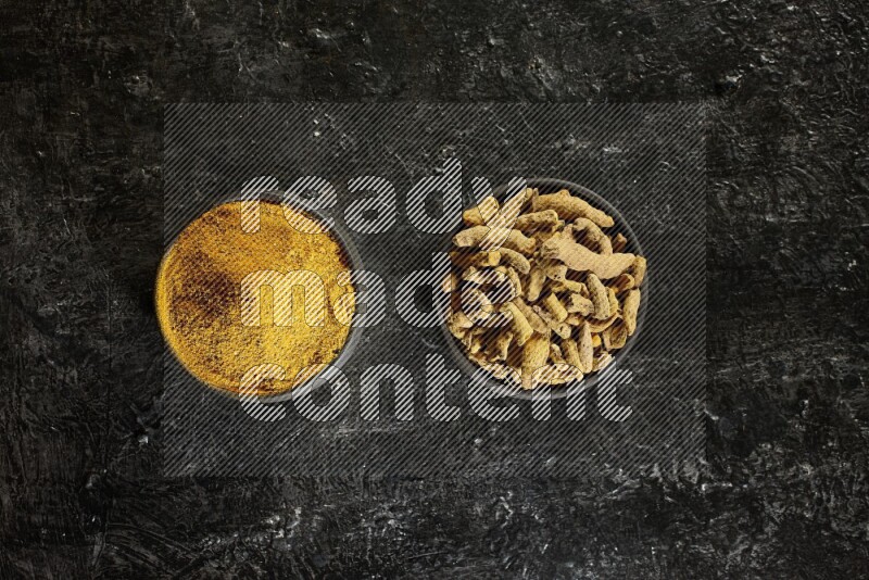 2 pottery black bowls, one full of turmeric powder and the other full of dried turmeric whole fingers on a textured black flooring