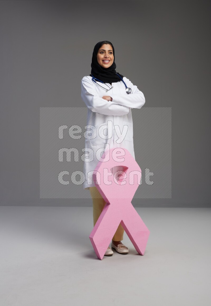 Saudi woman wearing lab coat with stethoscope standing holding awareness ribbon symbol standing on Gray background