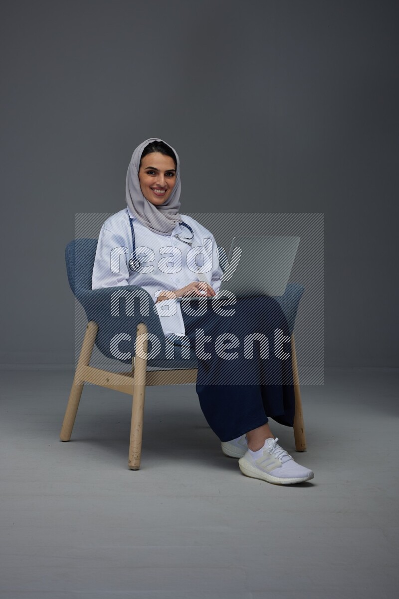 A doctor wearing a light gray head scarf sitting on a dark grey chair and using a laptop eye level on a grey background