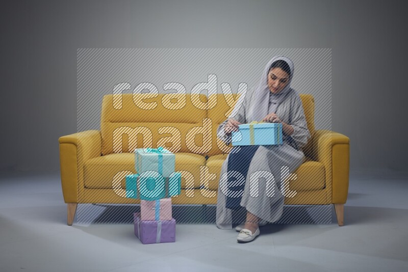 A Saudi woman wearing a light gray Abaya and head scarf sitting on a yellow sofa holding a gift box beside multi color gift boxes eye level on a grey background