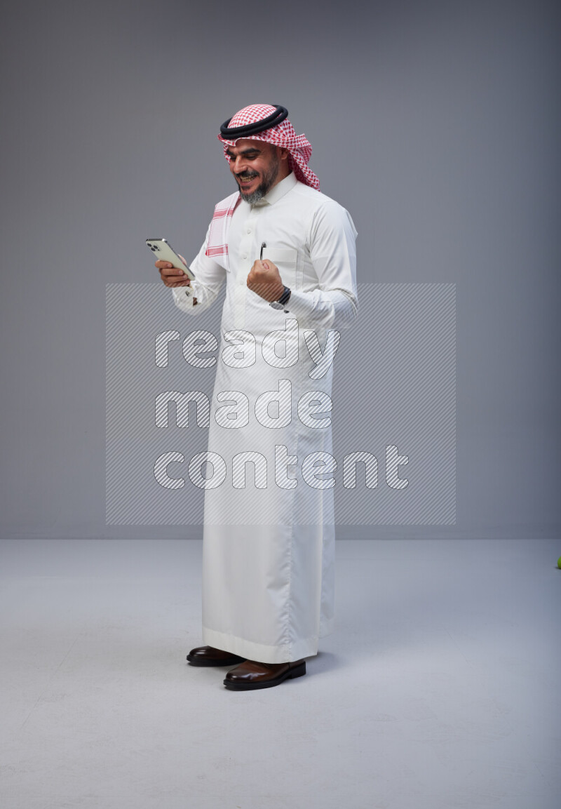 Saudi man Wearing Thob and red Shomag standing texting on phone on Gray background