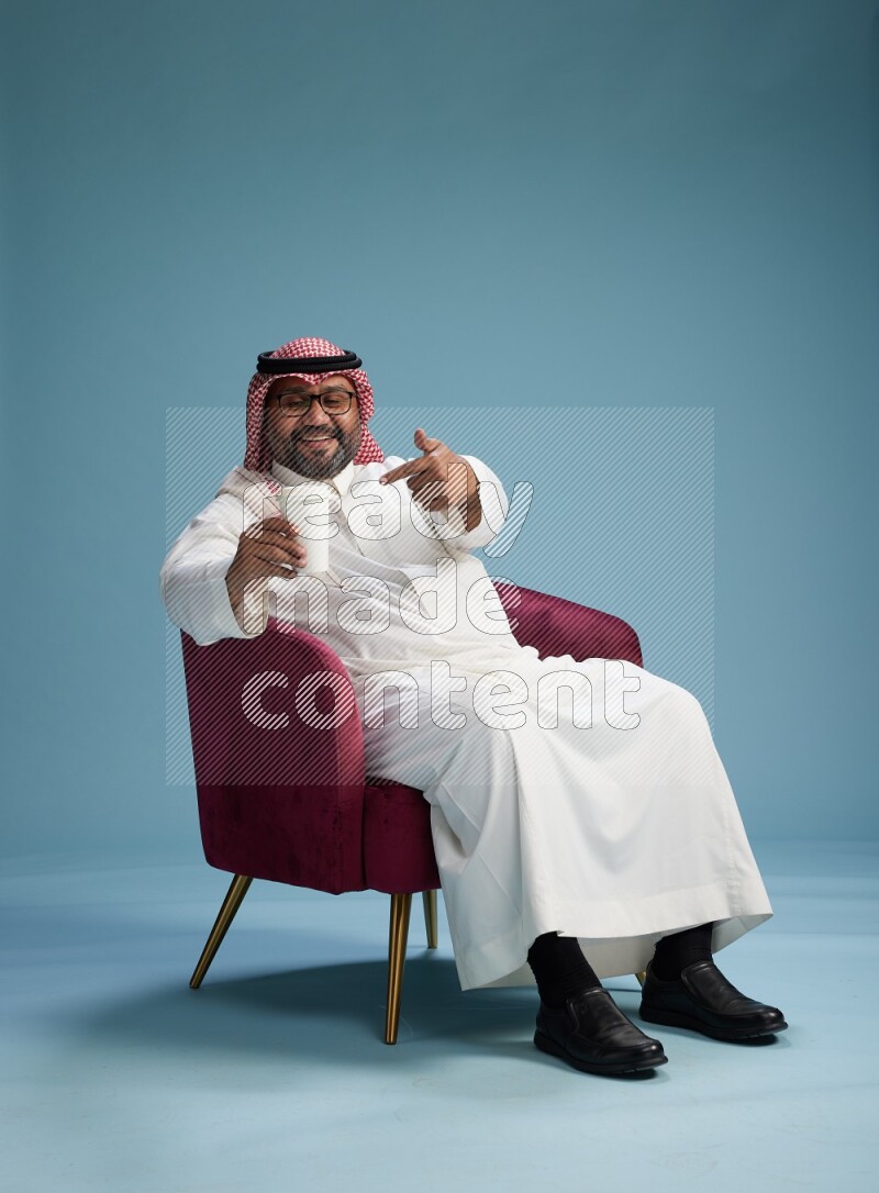 Saudi Man with shimag sitting on chair drinking coffee on blue background