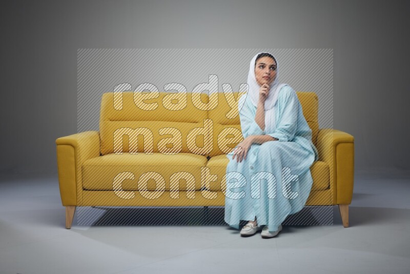 A Saudi woman wearing a light blue Abaya and white head scarf sitting on a yellow sofa and thinking and pointing up eye level on a grey background