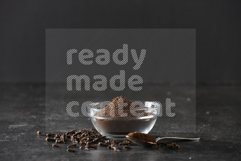 A glass bowl and a metal spoon full of cloves powder with gloves grains beside them on a textured black flooring