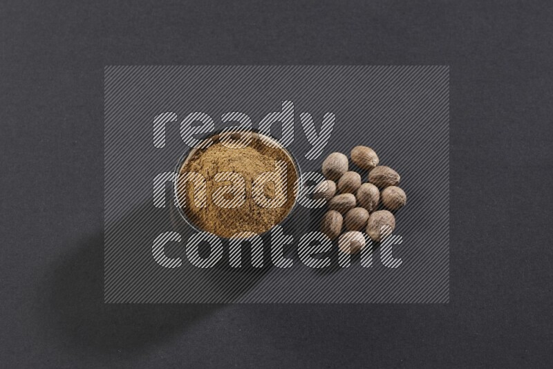 وعاء فخاري أسود ممتلئ ببودرة جوزة الطيب مع بعض حبوب جوزة الطيب بجانبه على خلفية سوداء