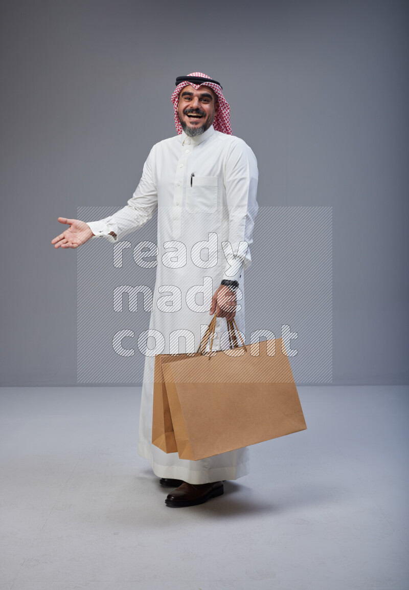 Saudi man Wearing Thob and red Shomag standing holding shopping bag on Gray background
