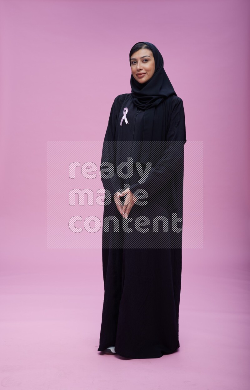 Saudi woman wearing pink ribbon on Abaya standing interacting with the camera on pink background