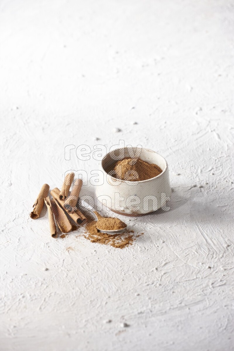 Ceramic beige bowl full of cinnamon powder and a metal spoon with cinnamon sticks next of it on a textured white background