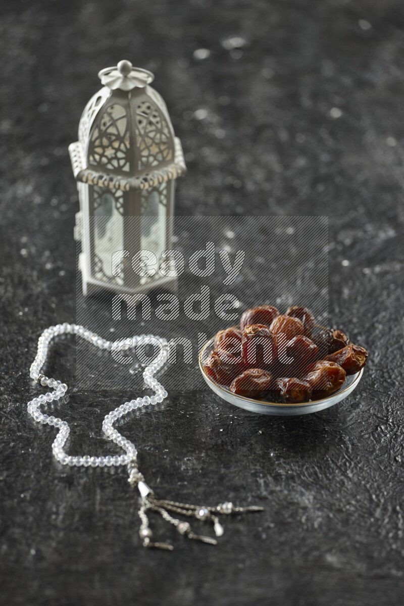 A white lantern with different drinks, dates, nuts, prayer beads and quran on textured black background