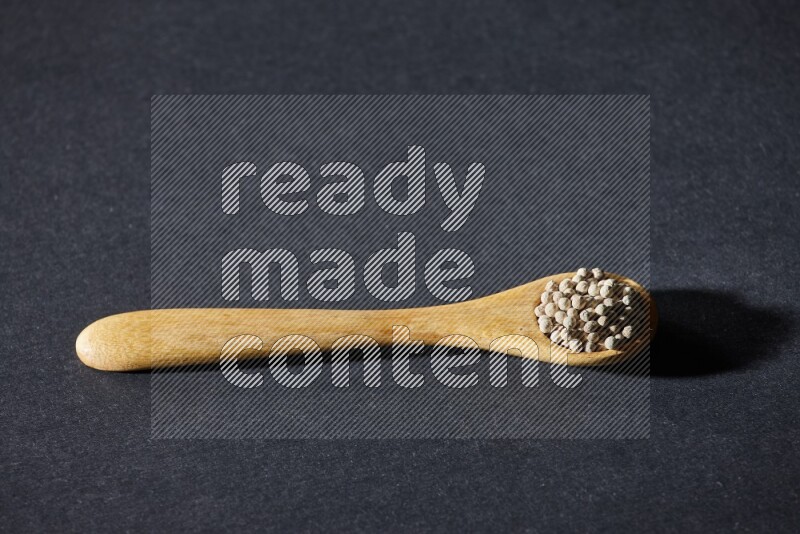 A wooden spoon full of white pepper beads on black flooring