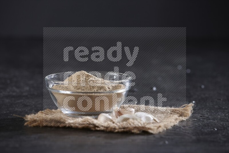 A glass bowl full of garlic powder placed on burlap fabric with garlic cloves on a textured black flooring