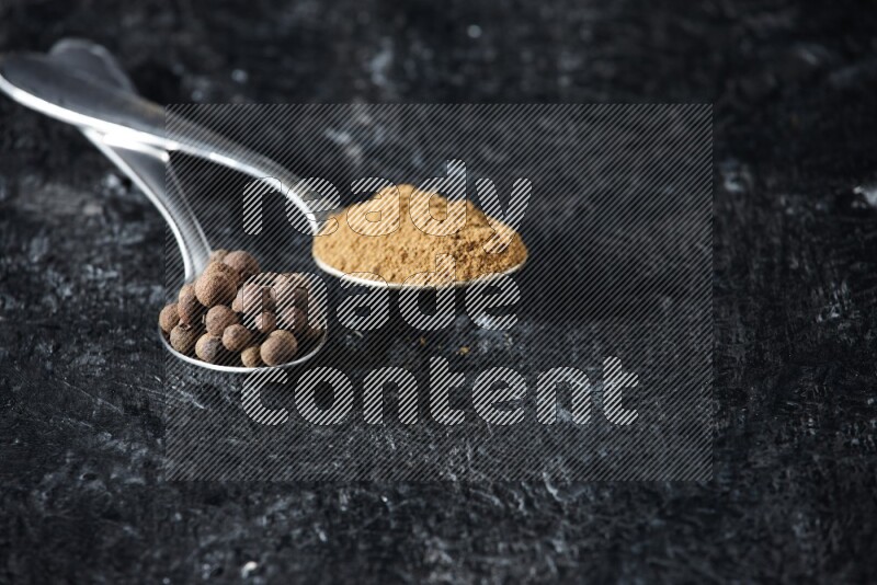 2 metal spoons full of allspice powder and whole balls on a textured black flooring