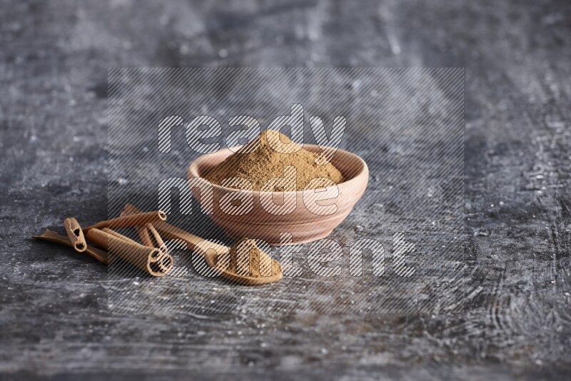 wooden bowl full of cinnamon powder and a wooden spoon full of it with cinnamon sticks on a textured black background