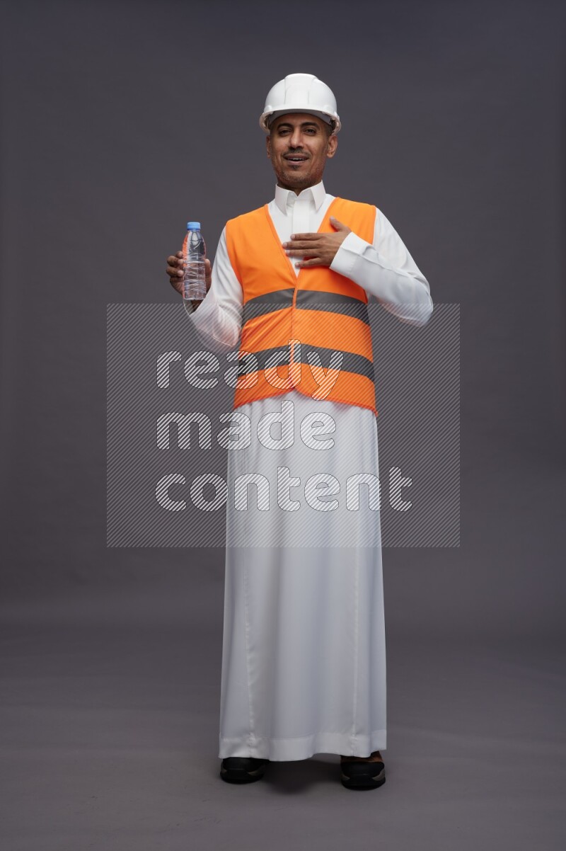 Saudi man wearing thob with engineer vest standing drinking water on gray background