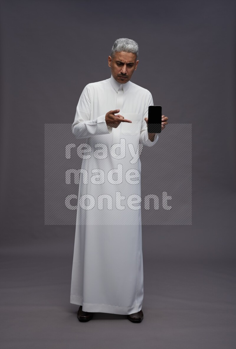 Saudi man wearing thob standing showing phone to camera on gray background