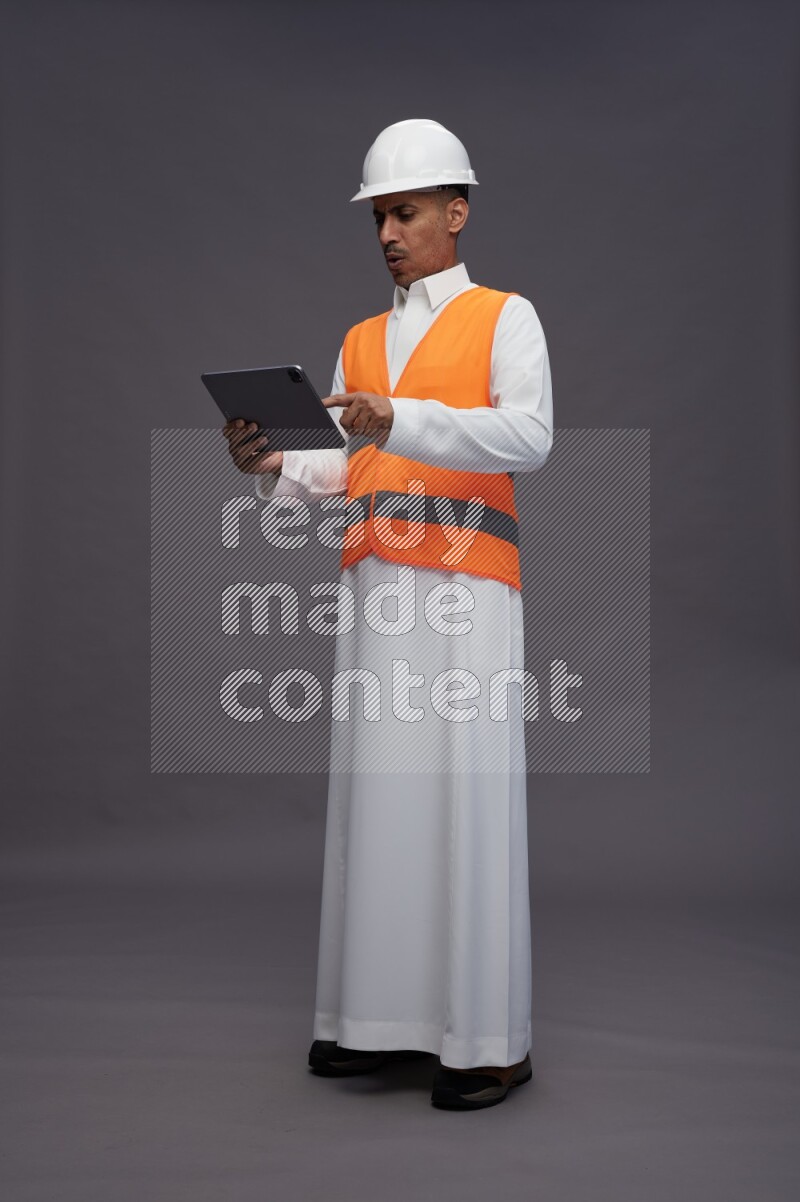 Saudi man wearing thob with engineer vest standing working on tablet on gray background