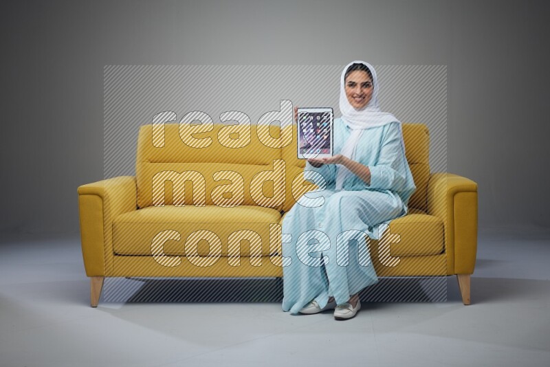A Saudi woman wearing a light blue Abaya and white head scarf sitting on a yellow sofa and showing her tablet's screen while pointing to it eye level on a grey background