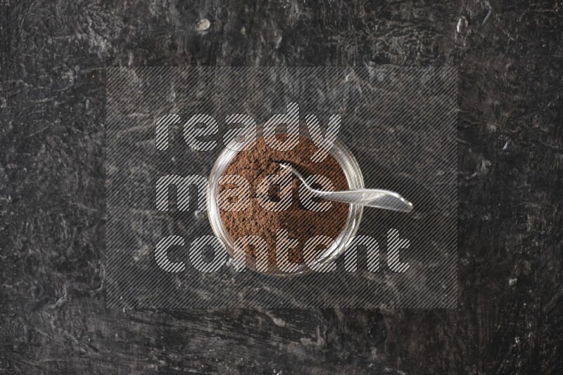 A glass jar full of cloves powder with a metal spoon on a textured black flooring