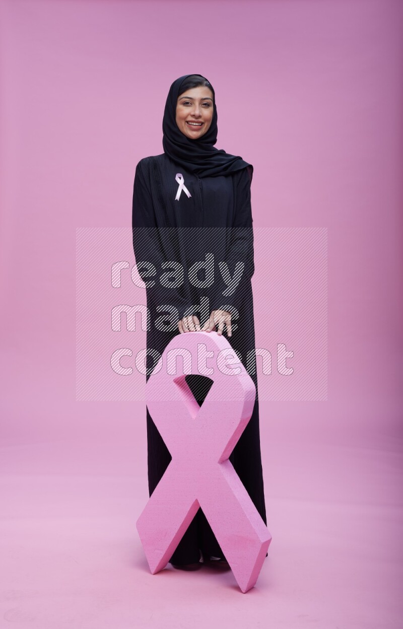Saudi woman wearing pink ribbon on abaya standing holding awareness ribbon on plain pink background