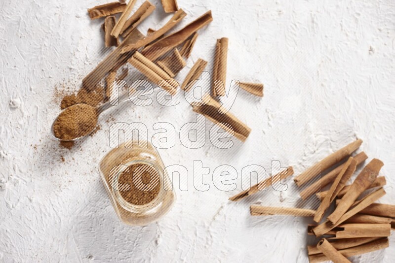 Herbal glass jar full cinnamon powder and a metal spoon surrounded by cinnamon sticks on a white background