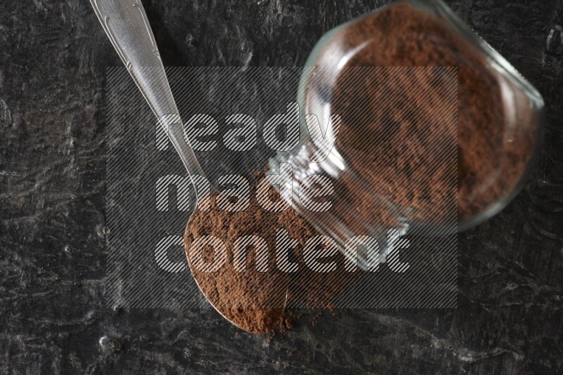A flipped glass spice jar and a metal spoon full of cloves powder on textured black flooring