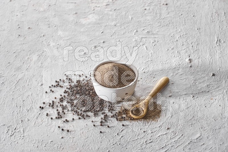 A pottery white bowl full of black pepper powder and wooden spoon full of powder and black pepper beads on a textured white flooring