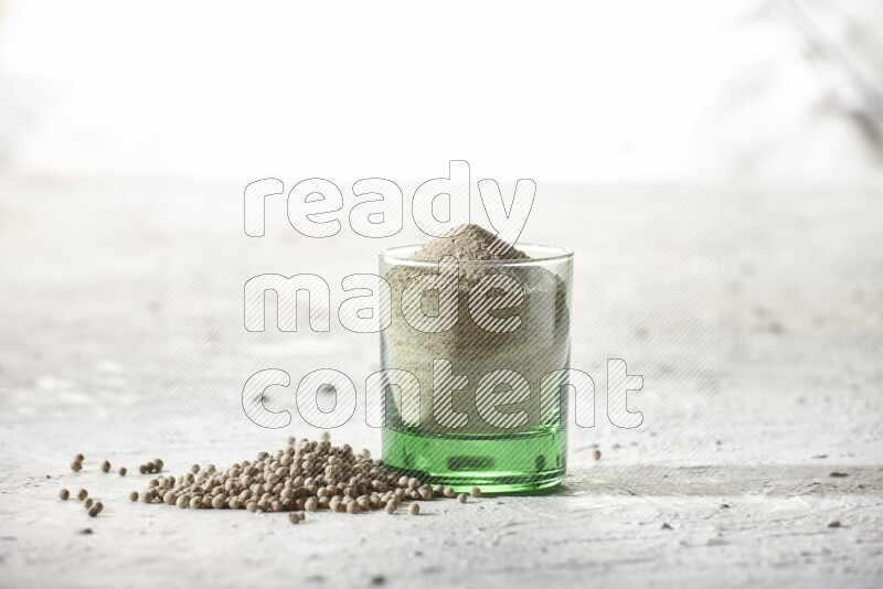 A green glass cup full of white pepper powder with white pepper beads on textured white flooring