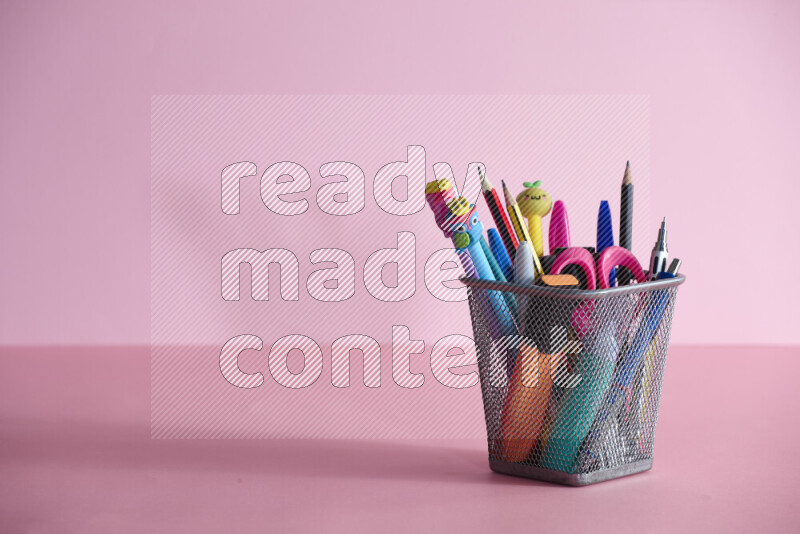 Pens, pencils and other different school supplies in a metal pen holder on rose background