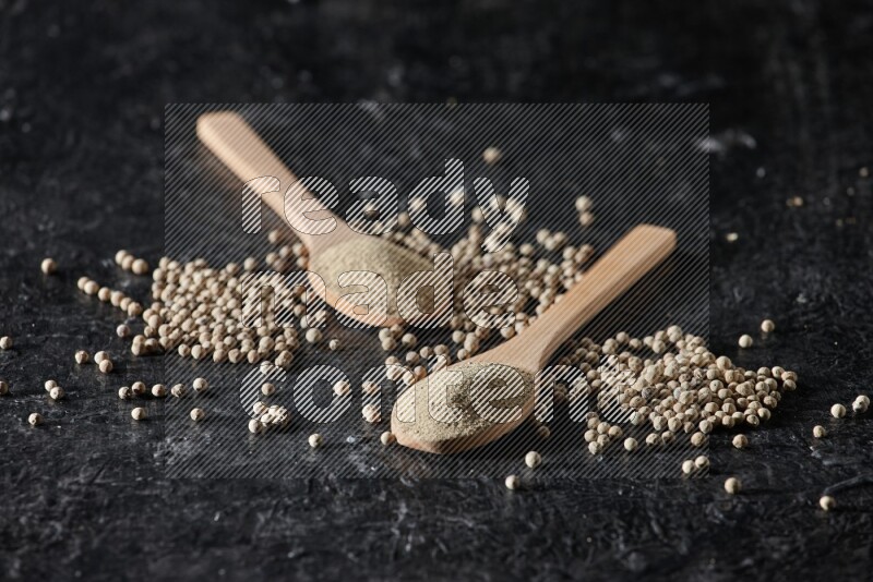 2 wooden spoons full of white pepper powder with white pepper beads on textured black flooring