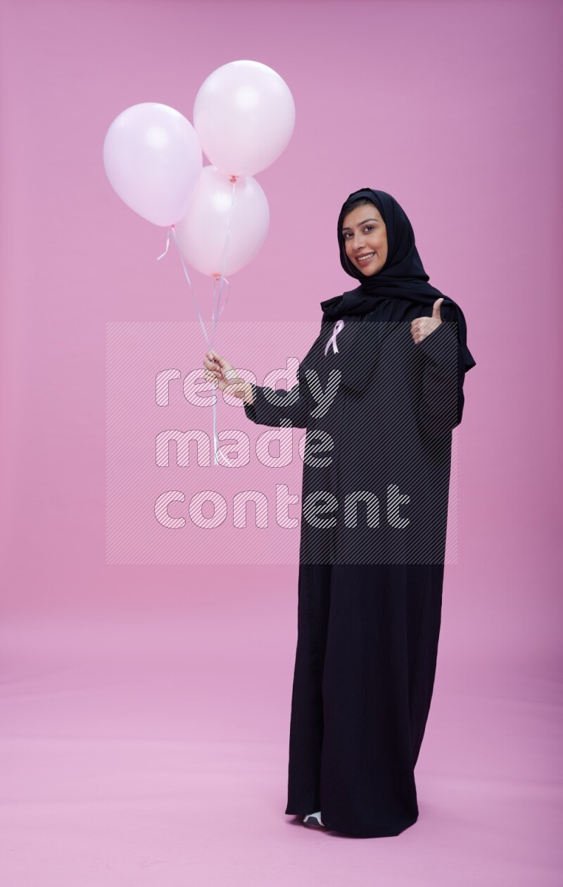 Saudi woman wearing pink ribbon on Abaya standing holding balloons on plain pink background