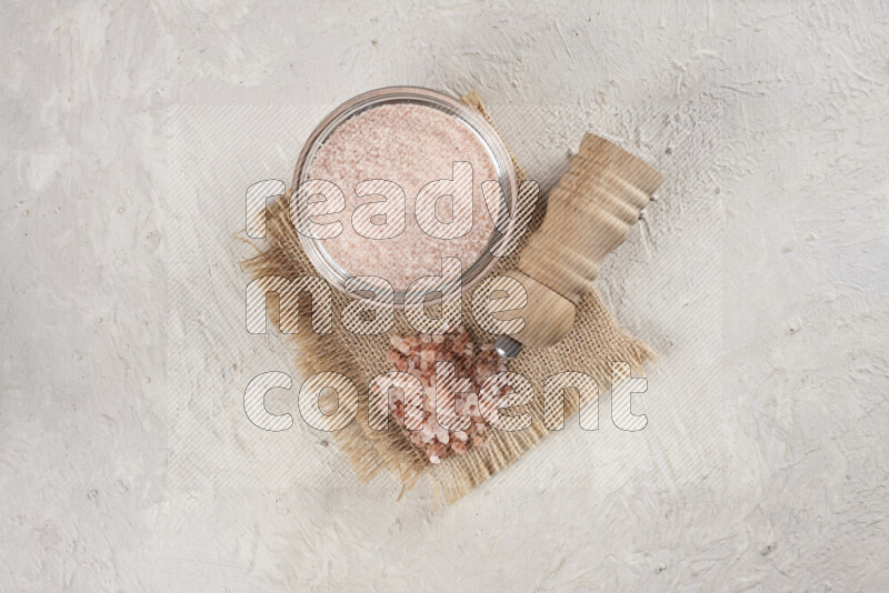 A glass bowl full of pink himalayan salt with a wooden grinder on a burlap fabric all on white background