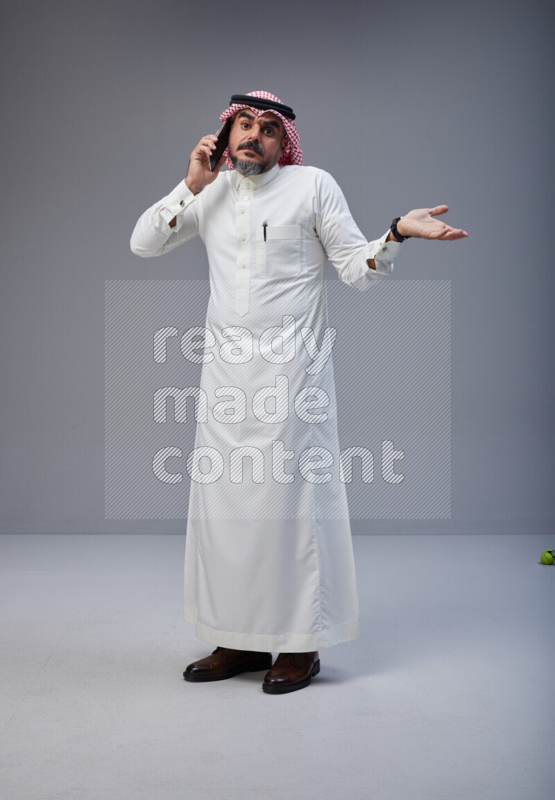 Saudi man Wearing Thob and red Shomag standing talking on phone on Gray background