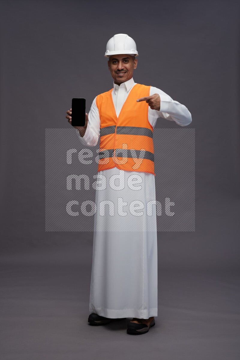Saudi man wearing thob with engineer vest standing showing phone to camera on gray background