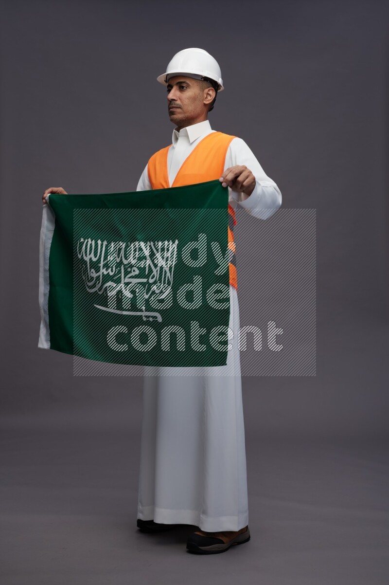 Saudi man wearing thob with engineer vest standing holding Saudi flag on gray background