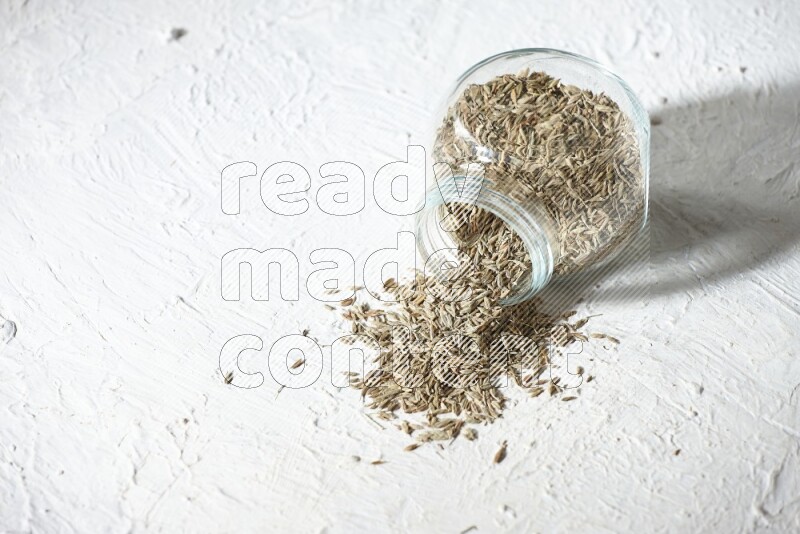 A flipped glass spice jar full of cumin seeds and the seeds spilled out on textured white flooring