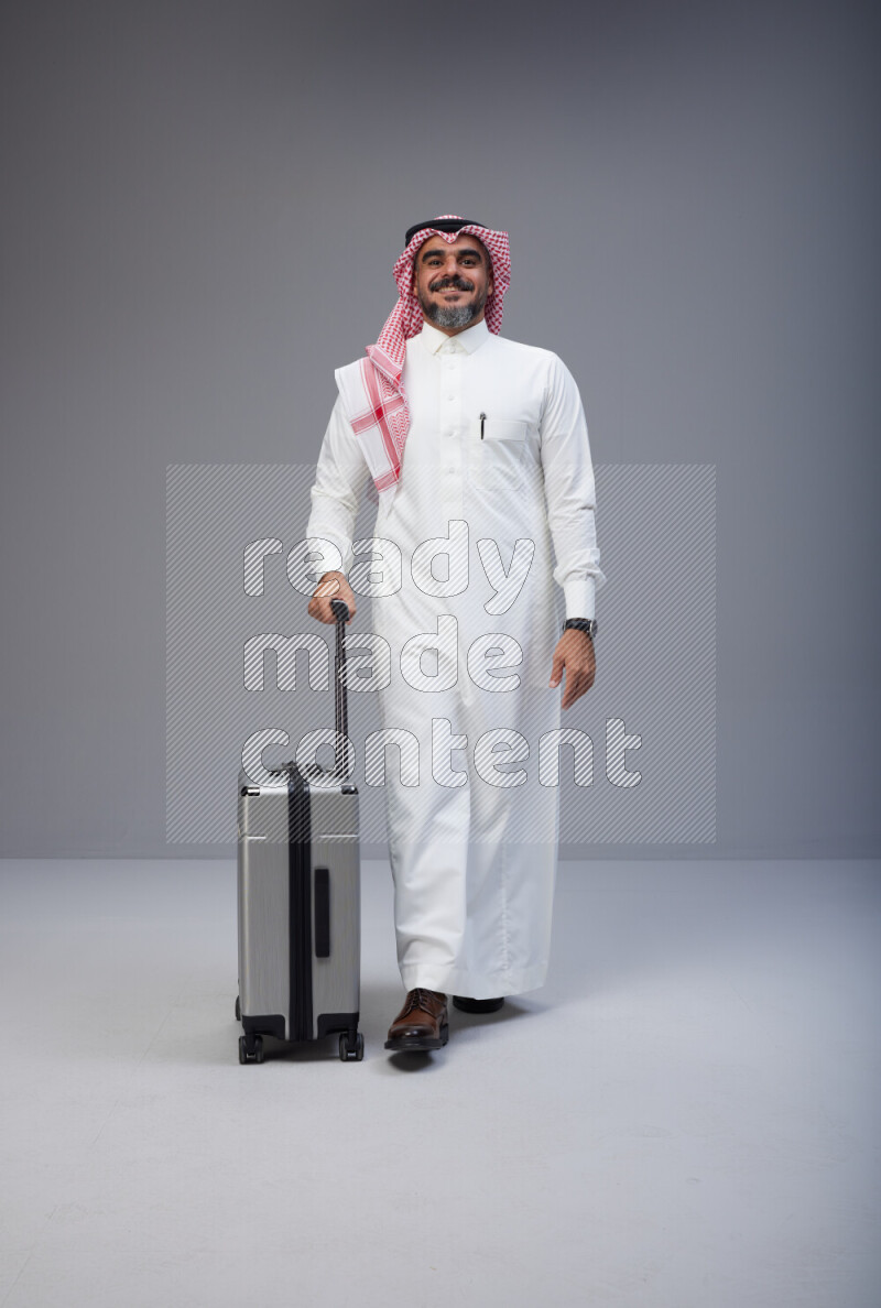 Saudi man wearing Thob and red Shomag standing holding Travel bag on Gray background