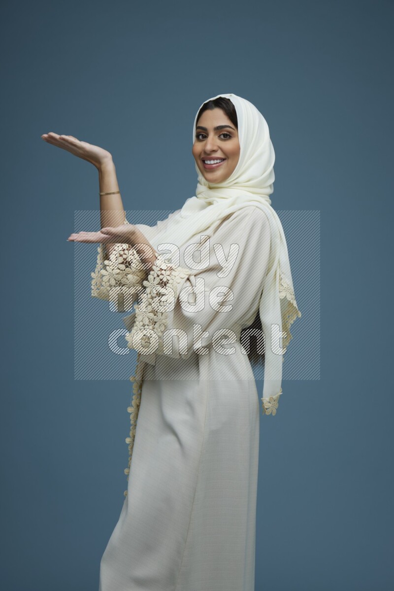A Saudi woman pointing in a blue background wearing an off-white Abaya Hijab