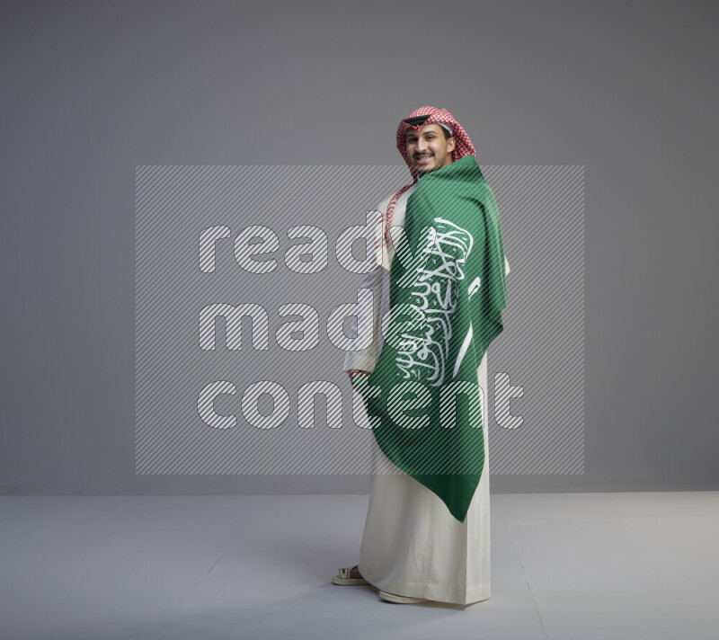 A saudi man standing wearing thob and red shomag holding big saudi flag on gray background