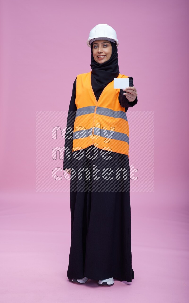Saudi woman wearing Abaya with engineer vest and helmet standing holding ATM card on pink background
