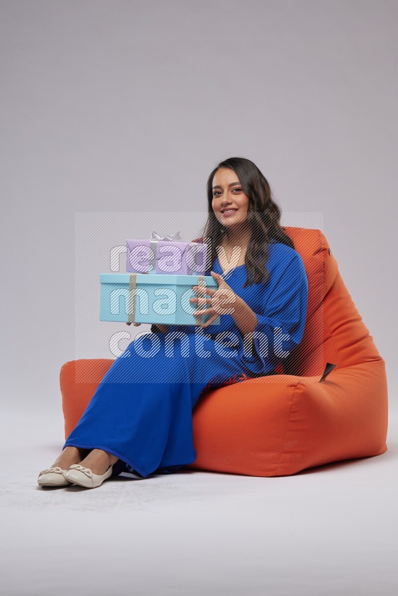 A woman sitting on an orange beanbag wearing Jalabeya holding a gift box