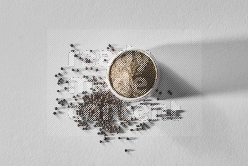 A white ceramic bowl full of black pepper powder and black pepper beads spread on white flooring